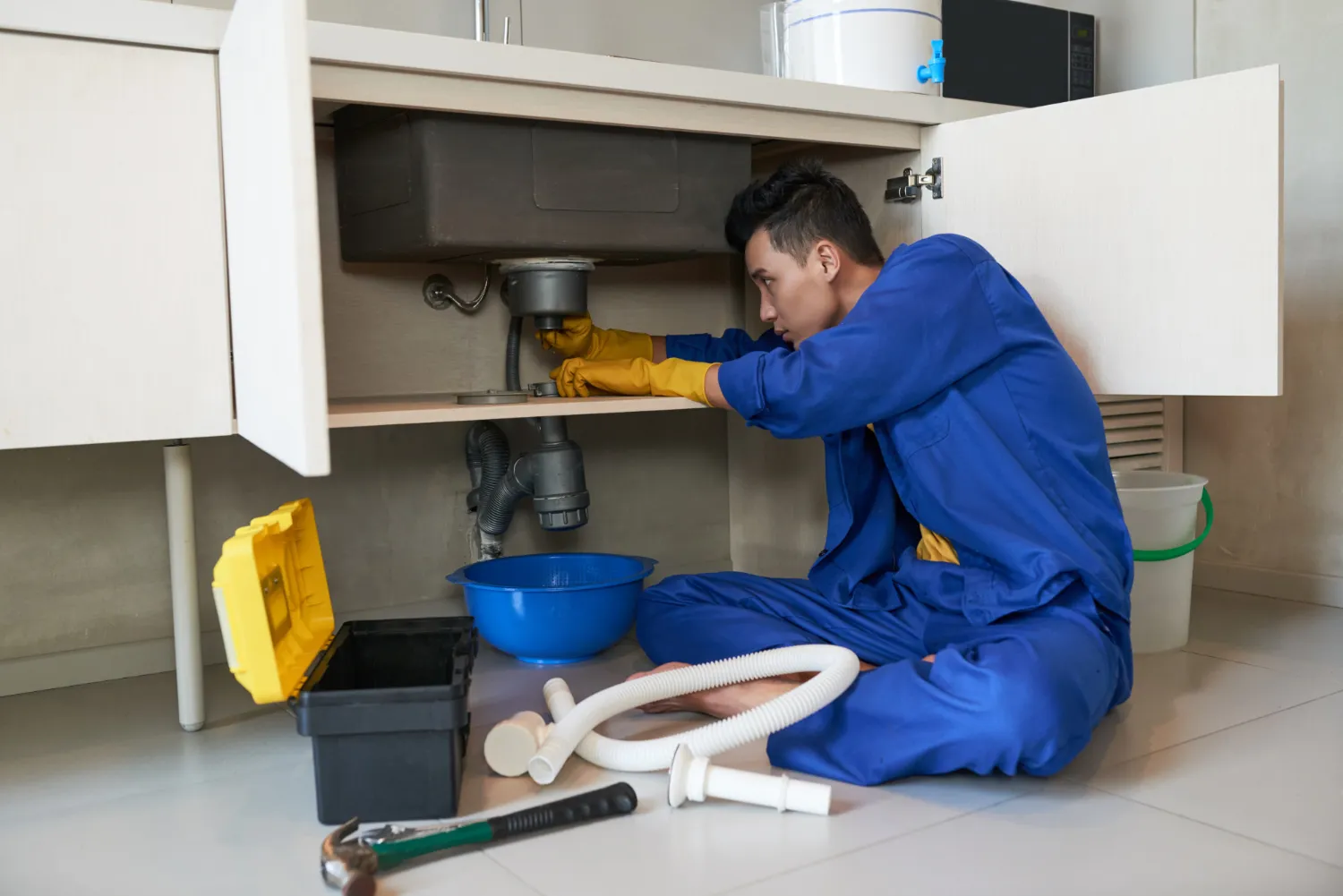 A plumber in blue overalls works under a kitchen sink, using tools and parts like hoses and containers for repairs