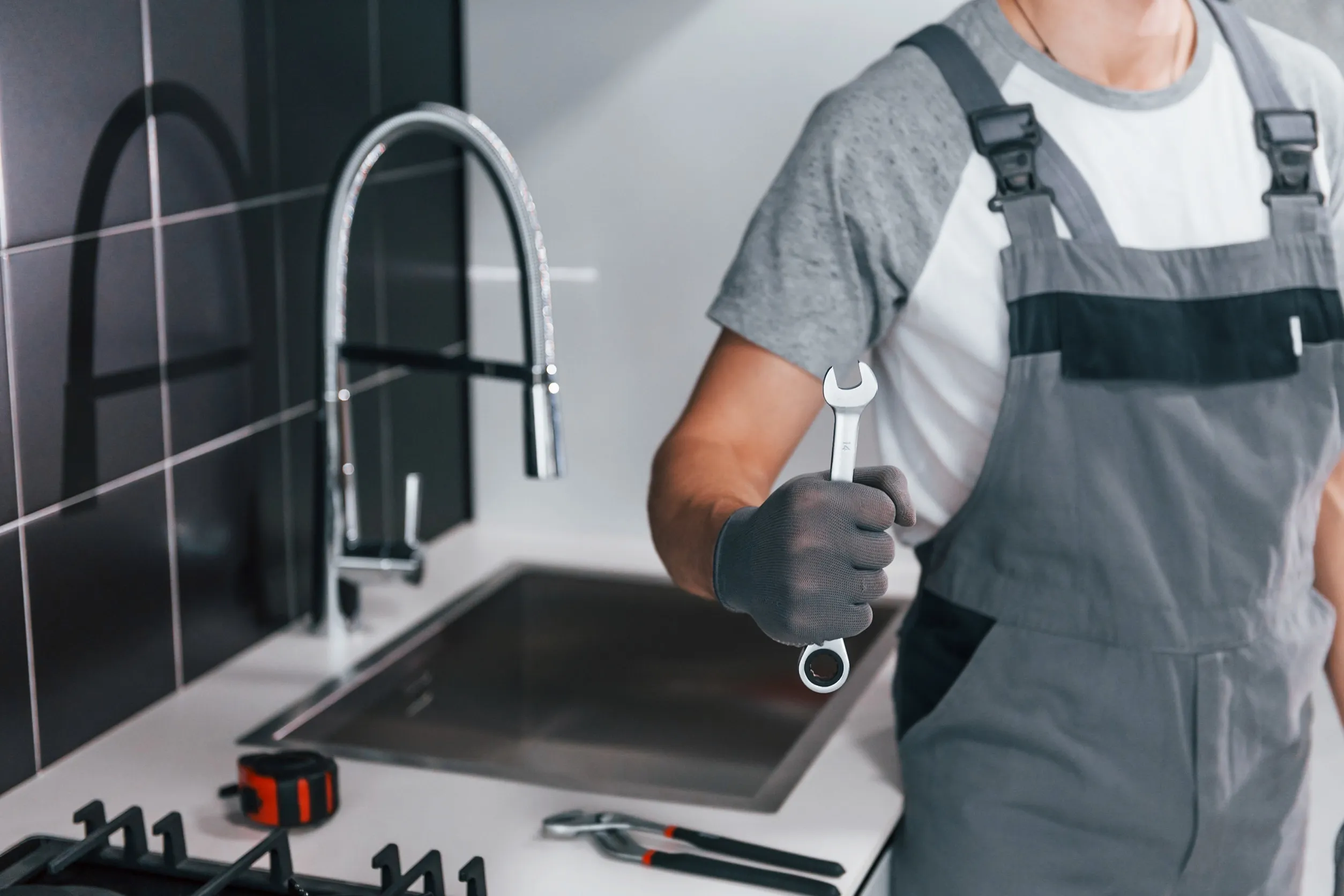 Close up view of young professional plumber in grey uniform holding wrench in hand on the kitchen